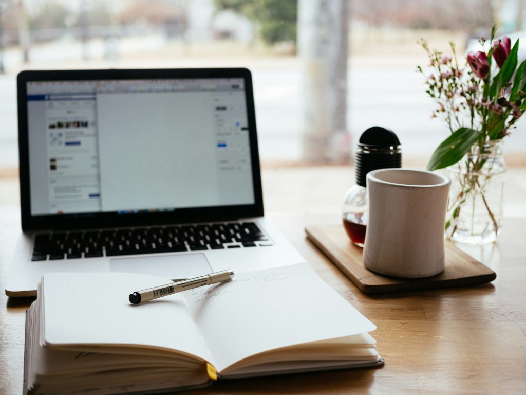 A laptop sits open next to a notebook and cup of coffee. Often the first steps in a budding journalist-creator's career – or day.